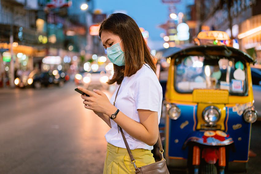 Una mujer joven con una máscara mira su teléfono mientras está en una calle de Bangkok